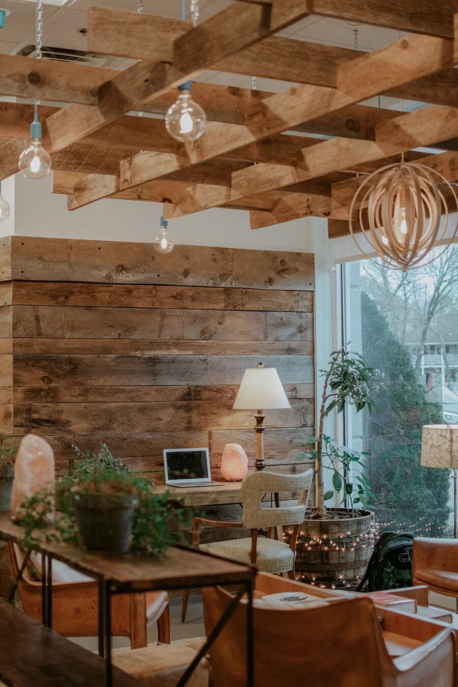 Un espace de travail chaleureux avec un plafond en poutres de bois apparentes, des suspensions lumineuses, des murs en bois rustique, un petit bureau avec un ordinateur portable et une lampe, des plantes en pot et de grandes fenêtres laissant entrer la lumière naturelle. L’espace offre une esthétique chaleureuse et organique avec des matériaux naturels.