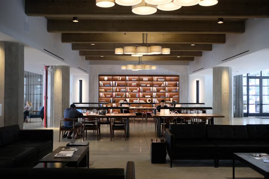 Grand espace de coworking avec tables communes, une grande bibliothèque murale et un éclairage moderne sous des poutres en bois.